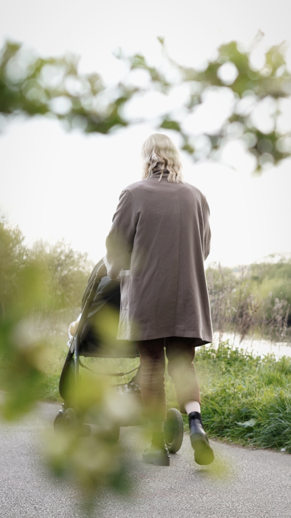 Amy is walking away from the camera pushing a pram, the foreground is framed by leaves